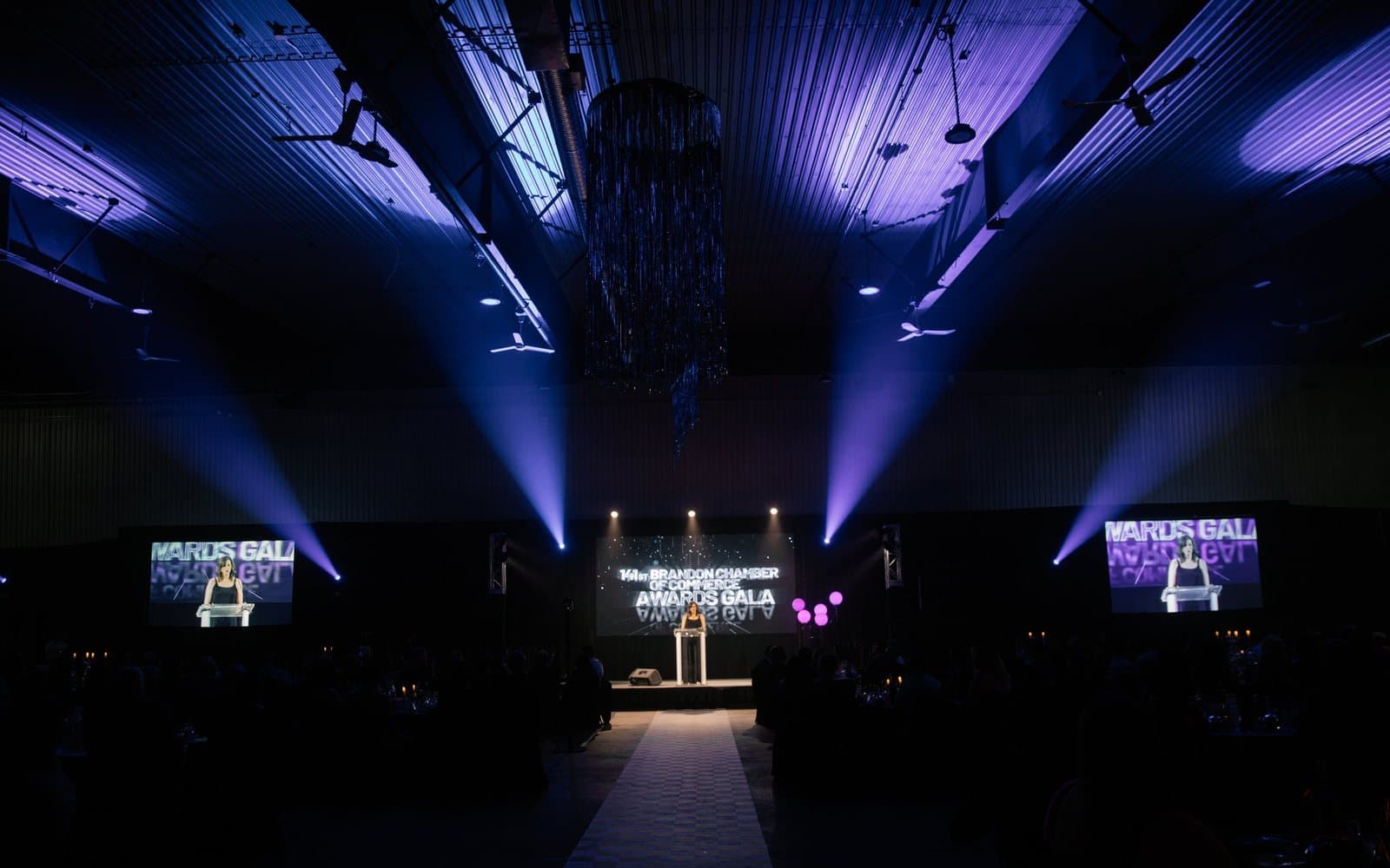The Brandon Chamber of Commerce Awards Gala stage under purple light, a speaker at the podium between three screens.