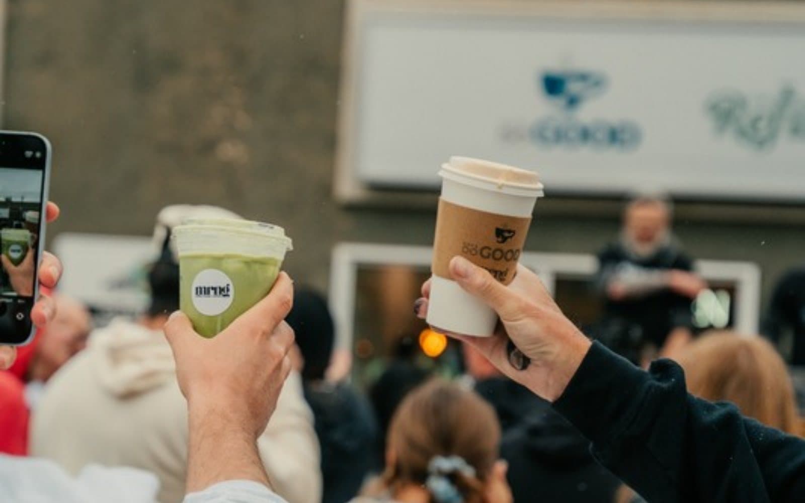 Two hands raise a coffee and a matcha with the mrng logo in front of a crowd at an outdoor morning event.